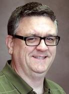 A man with short hair wearing glasses and a green collared shirt, smiling at the camera against a plain background.