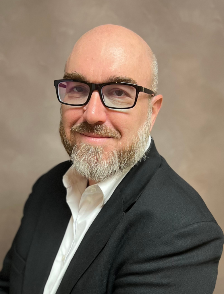 A bald man with glasses and a gray beard, wearing a black blazer and white shirt, poses against a plain background.