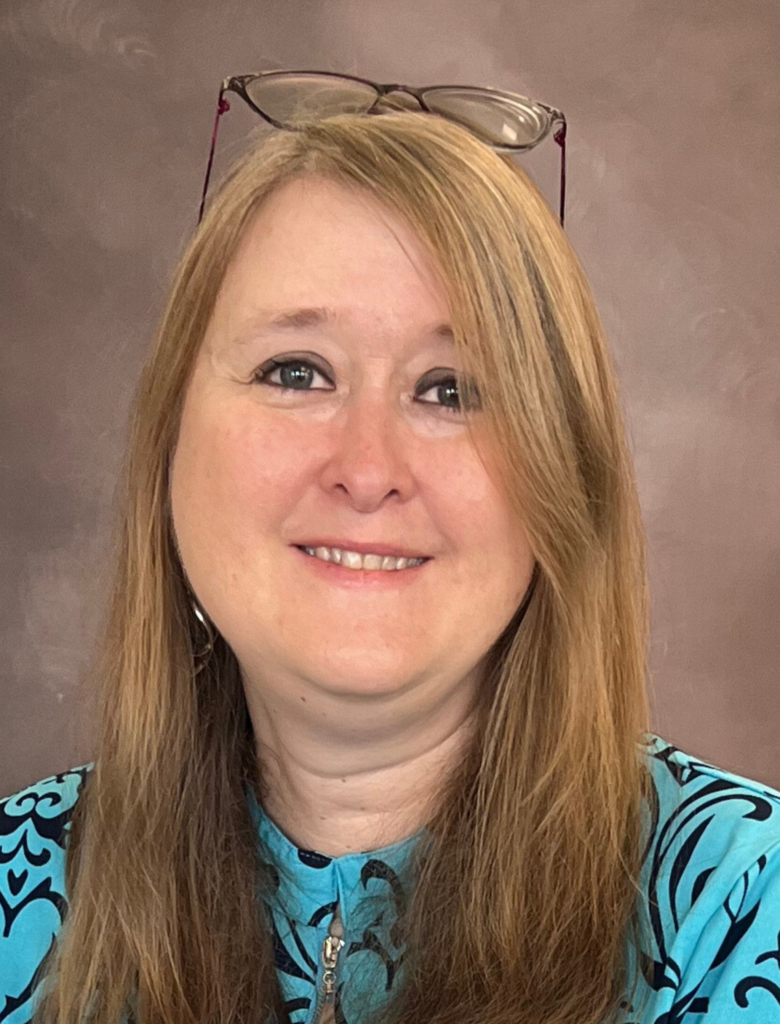 A woman with long, light brown hair smiles at the camera. She is wearing glasses on her head, a turquoise patterned top, and has a neutral brown background behind her.